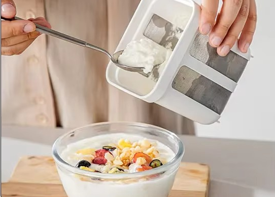 אישה אוחזת במסננת ויוגורט מסונן, ומוסיפה בכף גבינה רכה לקערת סלט פירות ויוגורט.

